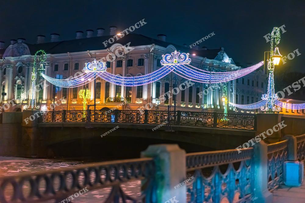 Фотография зимнего Санкт-Петербурга с видом на Невский проспект, украшенный рождественскими огнями и новогодними декорациями. На снимке показаны праздничные улицы исторического центра города, архитектура фасадов и отражения иллюминации на влажной брусчатке. Панорамный вид позволяет увидеть оживленное городское пространство и красоту зимнего Петербурга в период Рождества и Нового года. Изображение прекрасно передает атмосферу северной столицы России, праздничное настроение и сочетание городской архитектуры с праздничными украшениями. Фотография подходит для туристических публикаций, новогодних постеров, интерьерного оформления и коллекций зимних фотографий города.